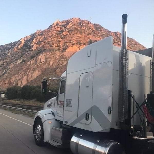 White semi-truck driving on a road with a mountain in the background. The mountain is rocky and appears to be illuminated by sunlight.