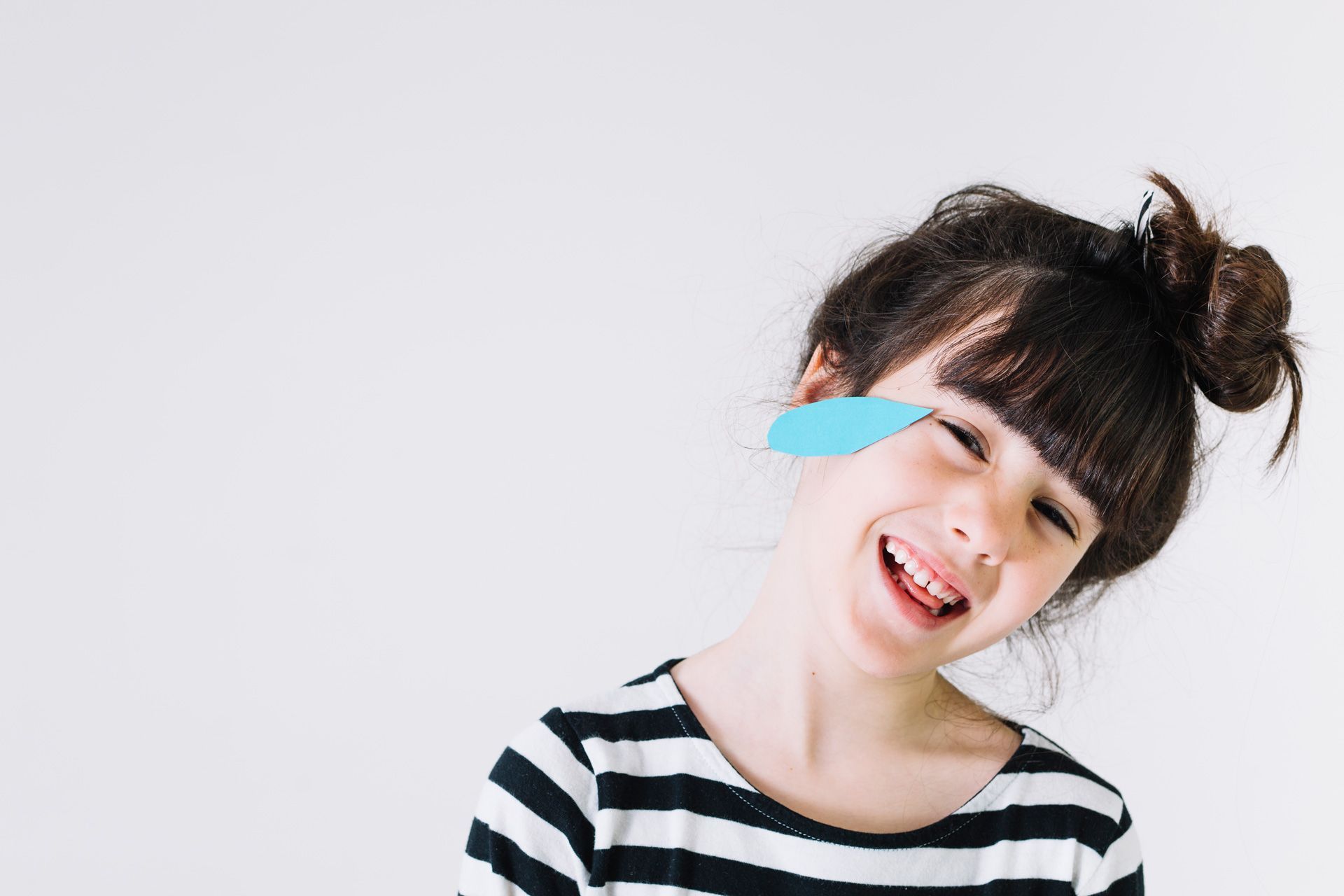 Girl with a paper feather over one eye, smiling broadly, wearing a striped shirt. Girl with a paper feather over one eye, smiling broadly, wearing a striped shirt.