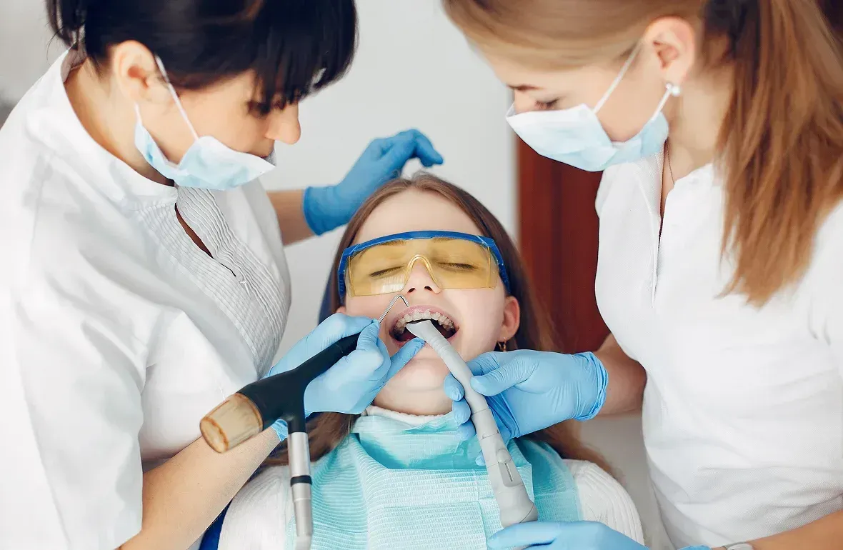 Dentist with two assistants examining a patient's teeth. The patient wears safety glasses, and all three are masked. Dentist with two assistants examining a patient's teeth. The patient wears safety glasses, and all three are masked.