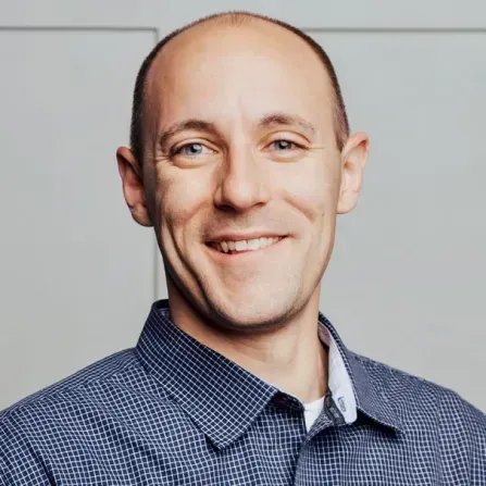 Smiling man with fair skin in a blue checkered shirt, against a light background.