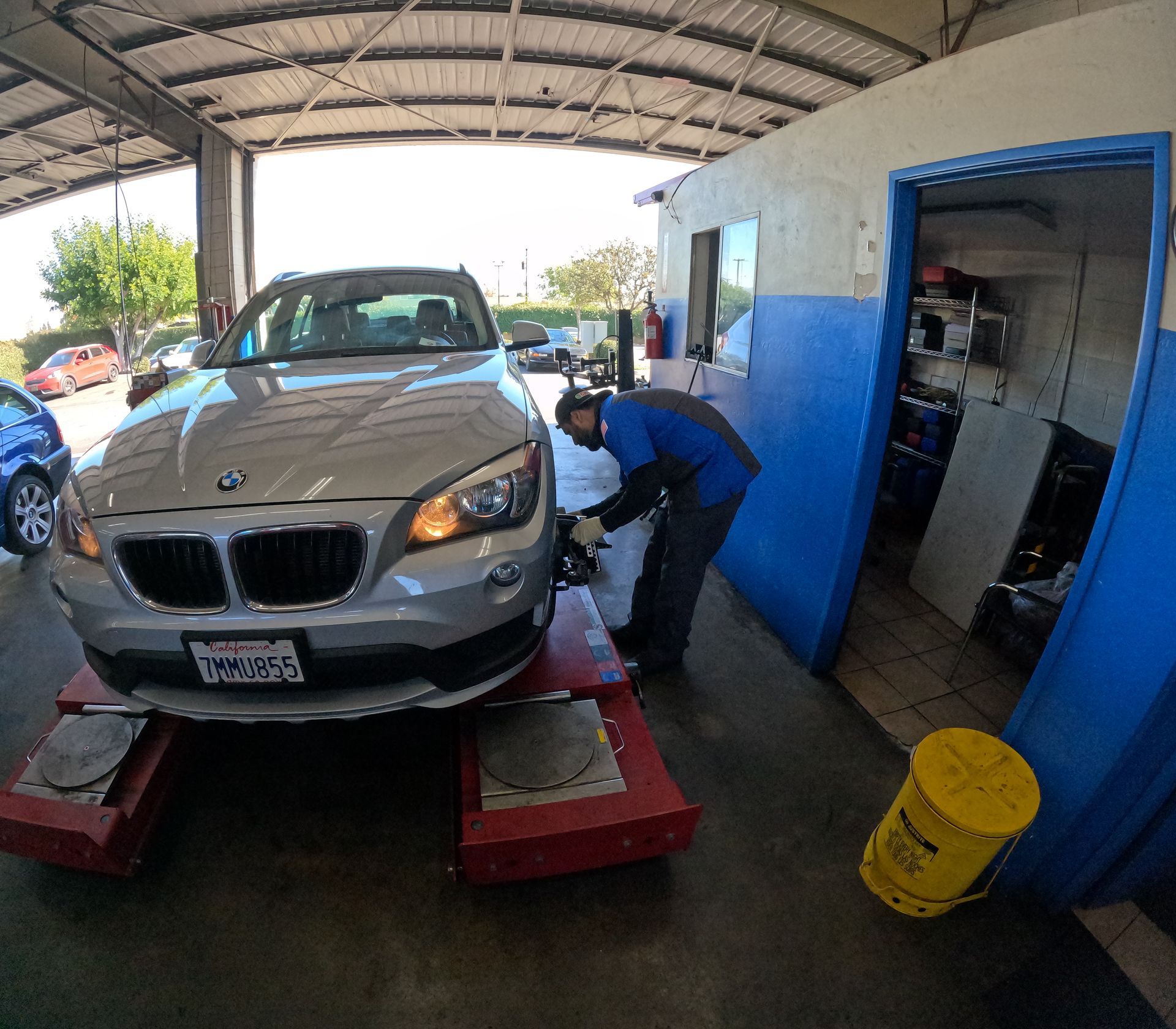 A mechanic working on a silver car at a garage. The car is on a lift. A yellow bucket is nearby.
