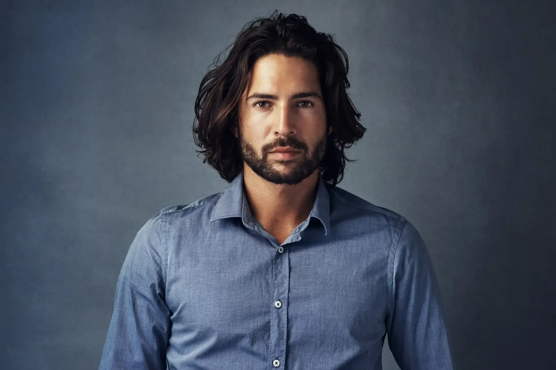 Portrait of a man with shoulder-length dark hair and beard, wearing a blue button-up shirt against a gray background