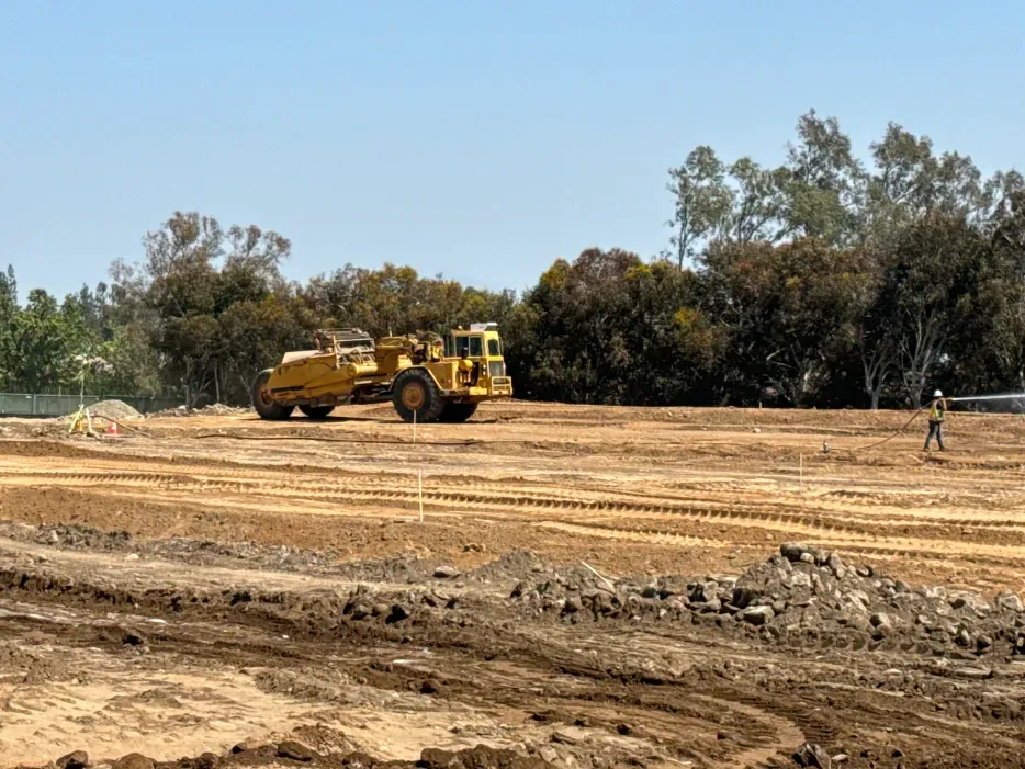 Construction site with a yellow scraper, leveling dirt. Person on the right. Trees and blue sky in the background.