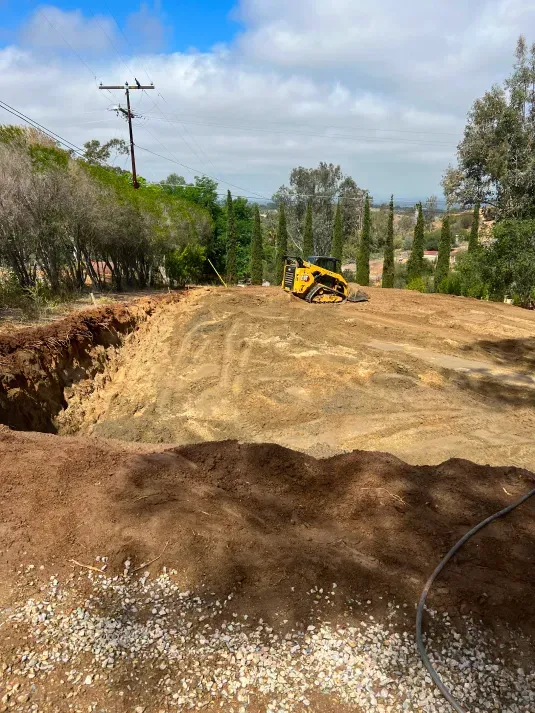 A yellow skid steer on a dirt lot with a large excavated area. Bright sunny day with a view in the distance.
