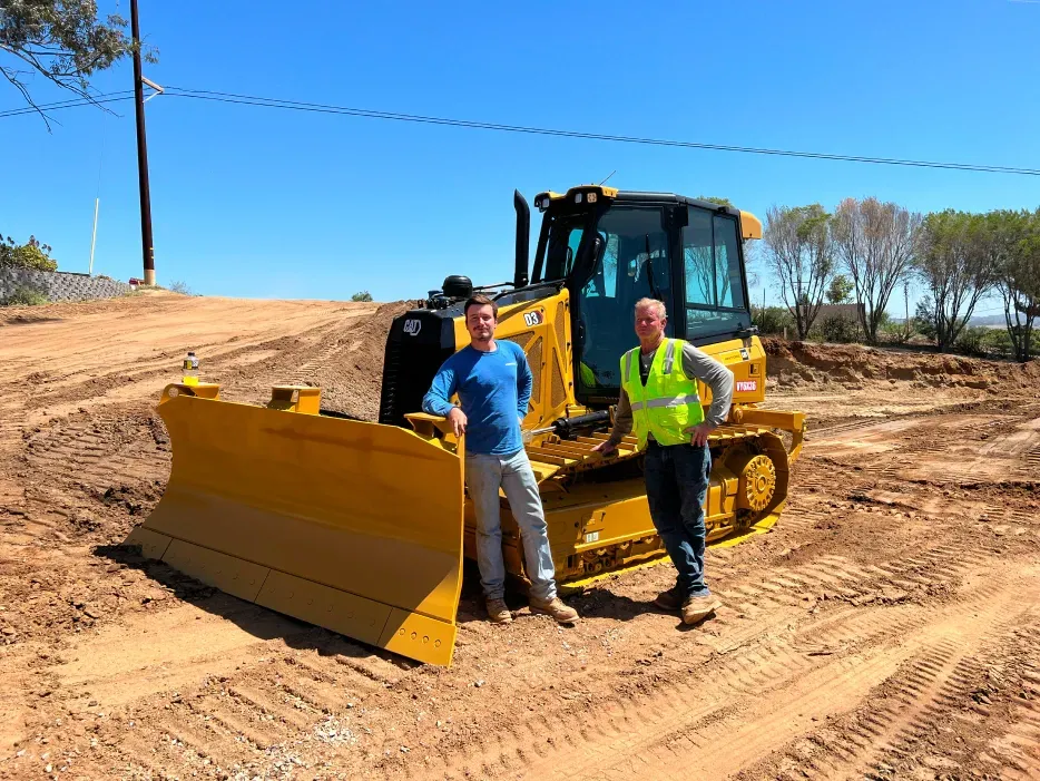 Two men stand by a yellow bulldozer in a dirt field on a sunny day.