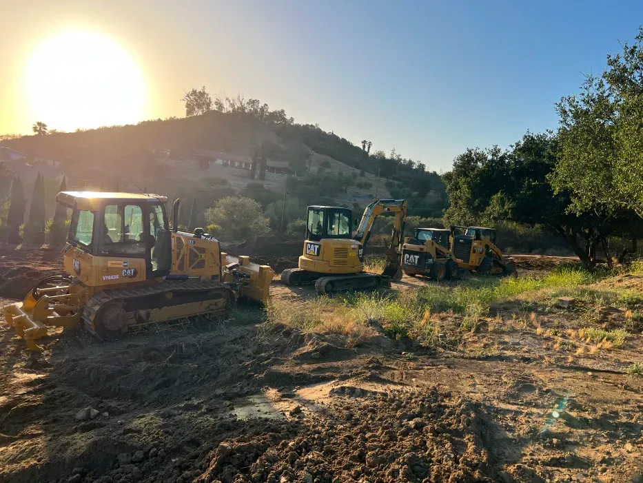 Construction site with heavy machinery, sunlight, and a hillside in the background.