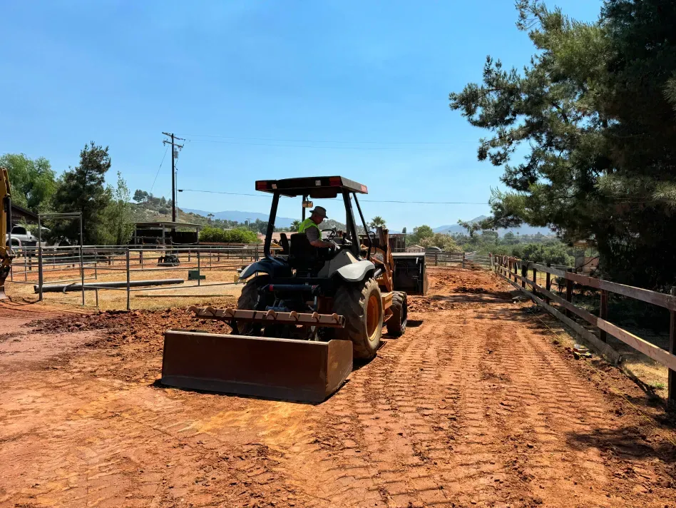 Tractor grading a dirt road in a sunny outdoor setting with fencing and trees.