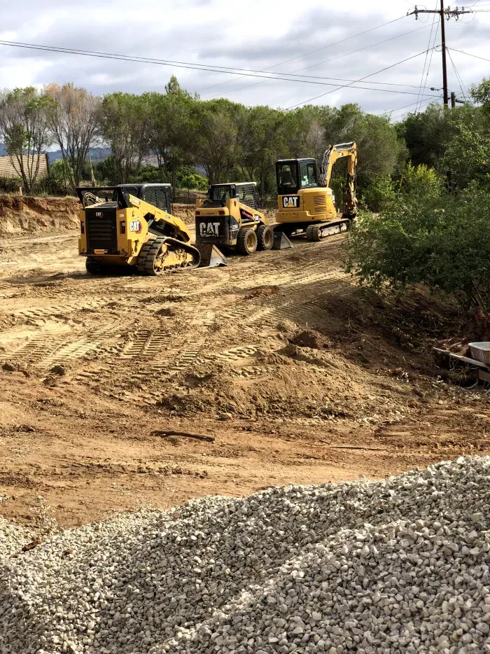 Construction site with three Caterpillar machines, digging in dirt and gravel; trees and sky in the background.