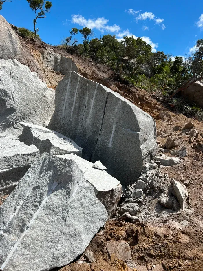 Large, gray rock face with carved lines, broken into large pieces, on a dirt hillside with trees under a blue sky.
