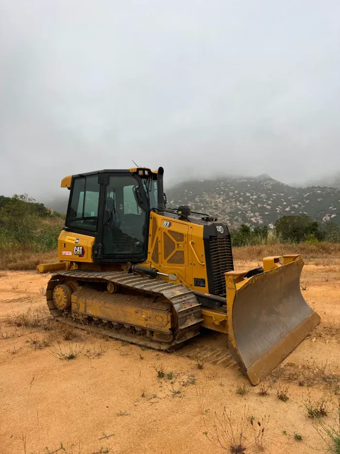 Yellow bulldozer on a dirt lot under a cloudy sky, with a hillside in the background.