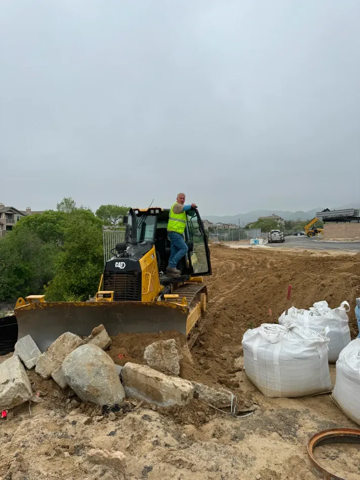 A construction worker standing on a yellow bulldozer, on a cloudy day, near large bags of debris and dirt.