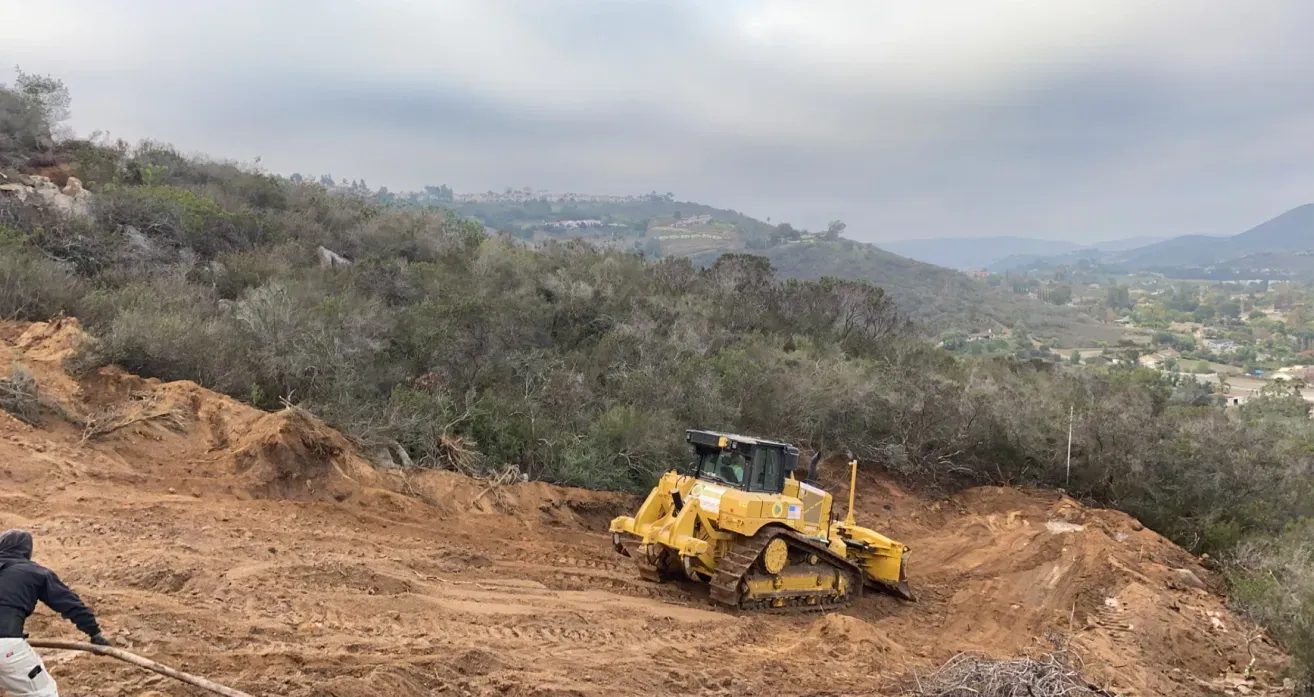 A bulldozer clearing land on a hillside under an overcast sky. A person works with a shovel.