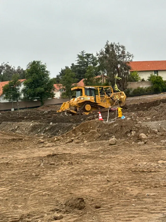 Yellow bulldozer on dirt hill at a construction site, houses and trees in the background.