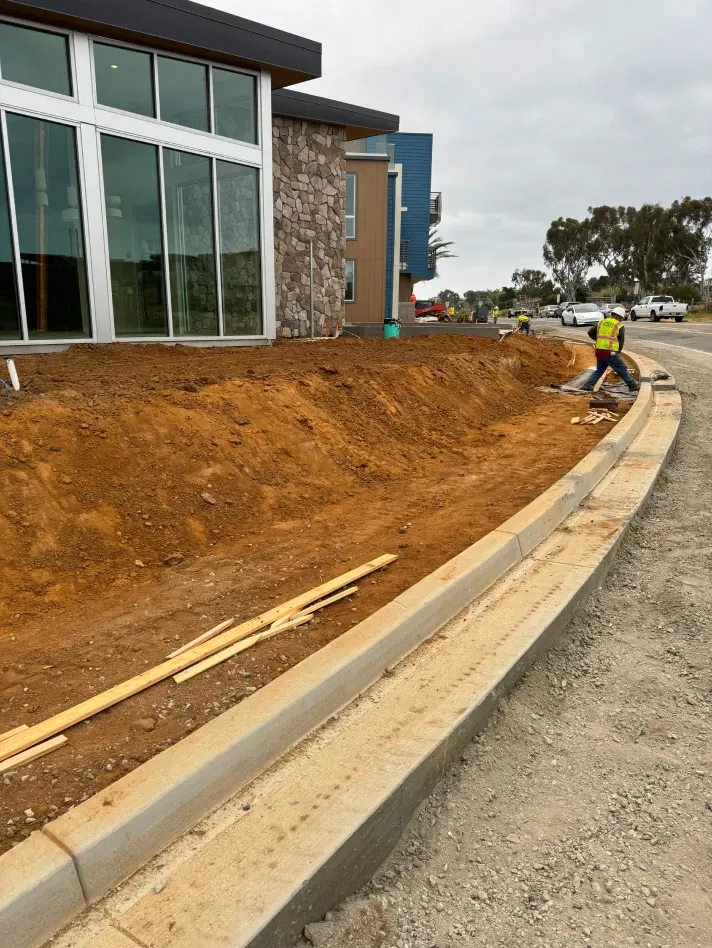 Construction site: Curb, dirt mound, building with windows, worker in safety vest.