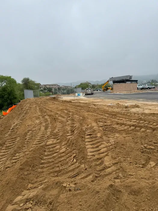 Construction site with dirt pile, excavator, and distant buildings under overcast sky.