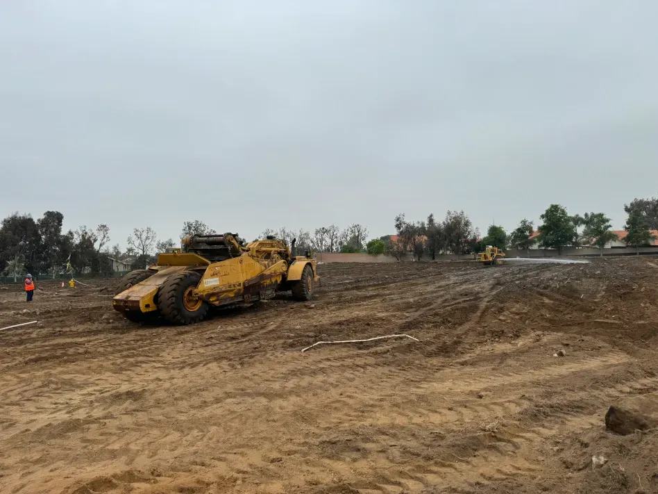 Heavy machinery leveling a muddy field on an overcast day; trees are in the background.