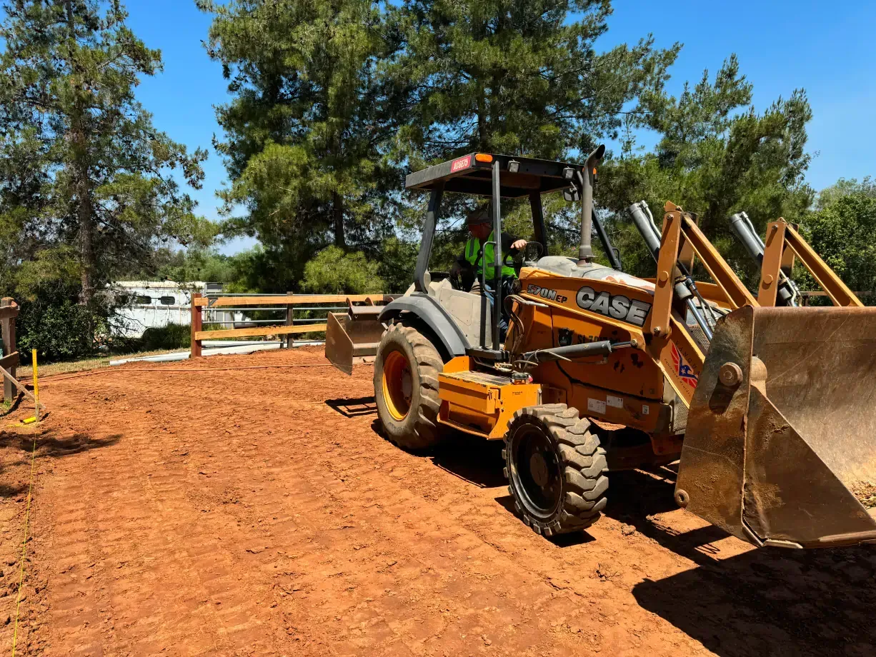 Backhoe on reddish-brown soil, preparing a site near a wooden fence under a blue sky.