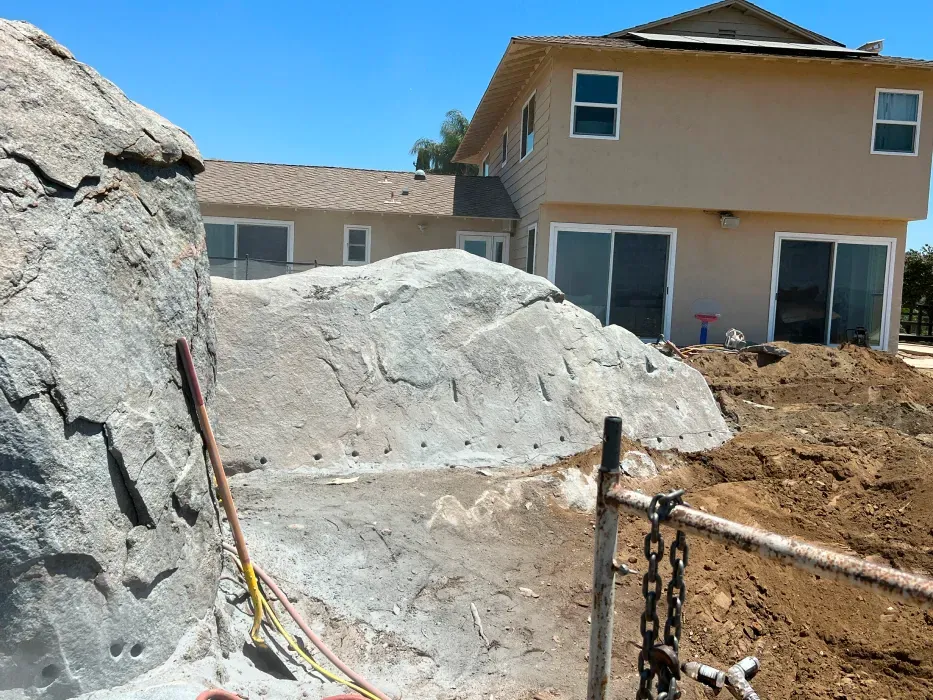 Construction site with large faux rock formations near a light brown house, sunny day.
