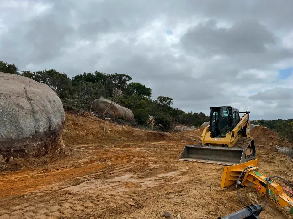 A yellow skid-steer loader on a dirt road next to large rocks and bushes, under a cloudy sky.