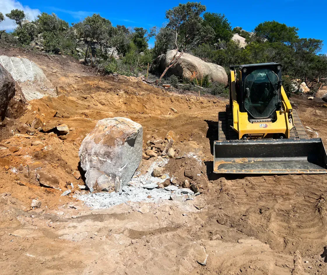 Yellow skid steer next to a large rock in a dirt excavation site under a blue sky.