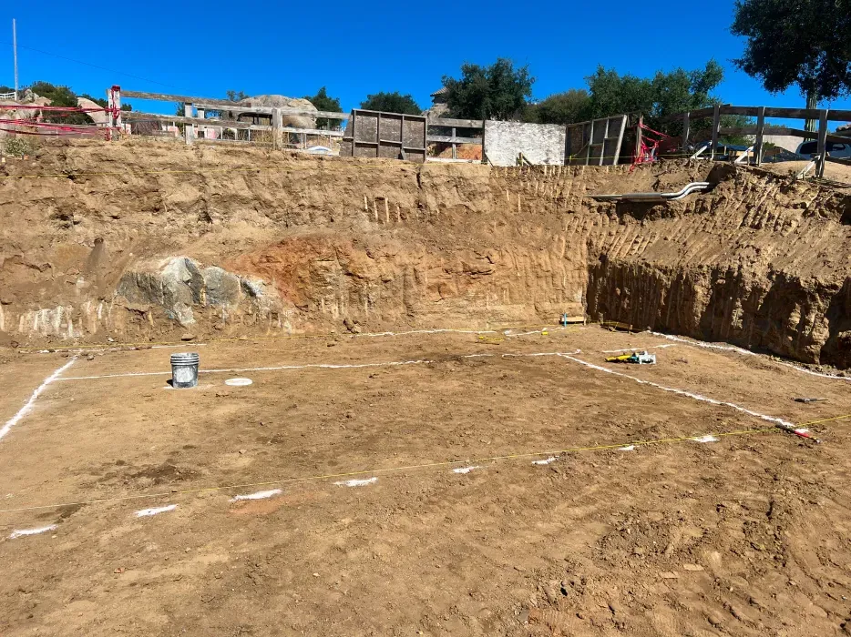 Construction site; large excavated area with white lines indicating foundation, dirt walls. Blue sky.