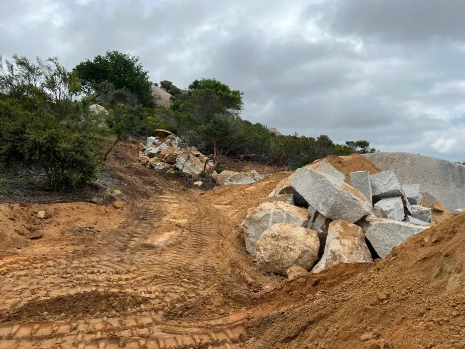Rocky terrain with boulders and a pile of cut stone; trees line the hillside, overcast sky.