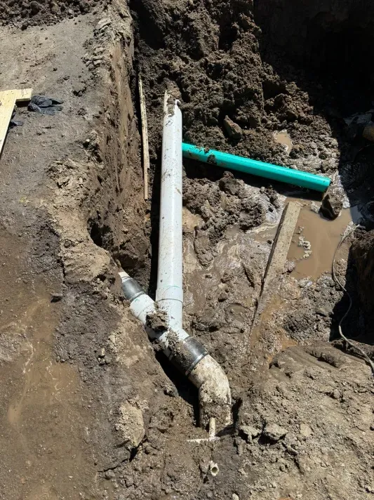 Excavated earth reveals plumbing pipes. Grey and black pipes are connected, with a green pipe nearby in a dirt trench.