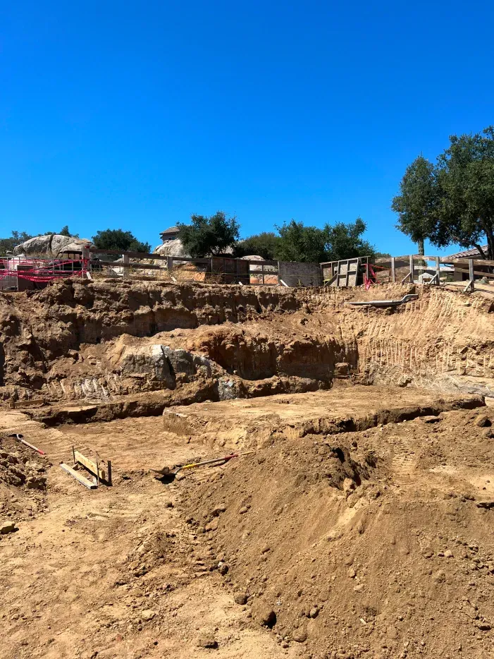 Construction site, earth and rocks excavated, clear blue sky, trees, and fencing in the background.
