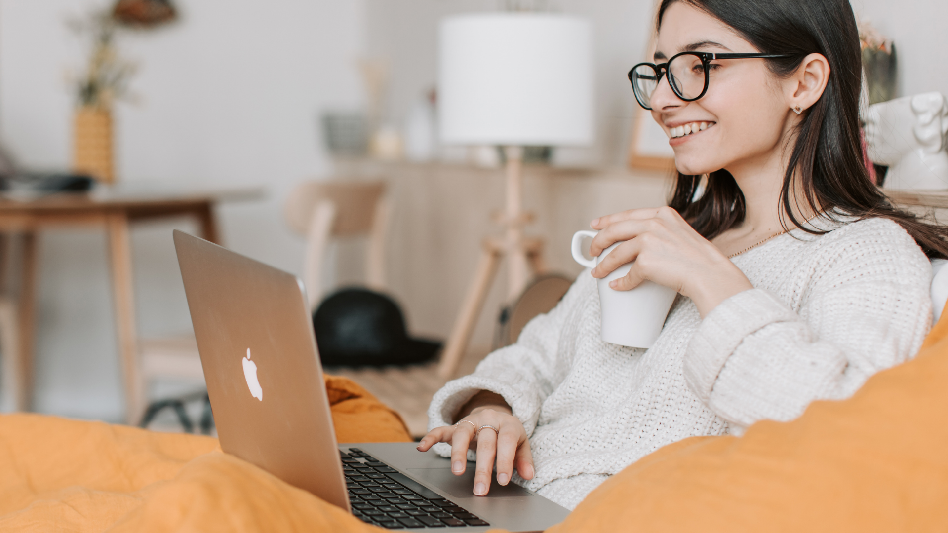 A person in a cream-colored sweater relaxes in bed with a laptop and a mug, smiling at the screen.