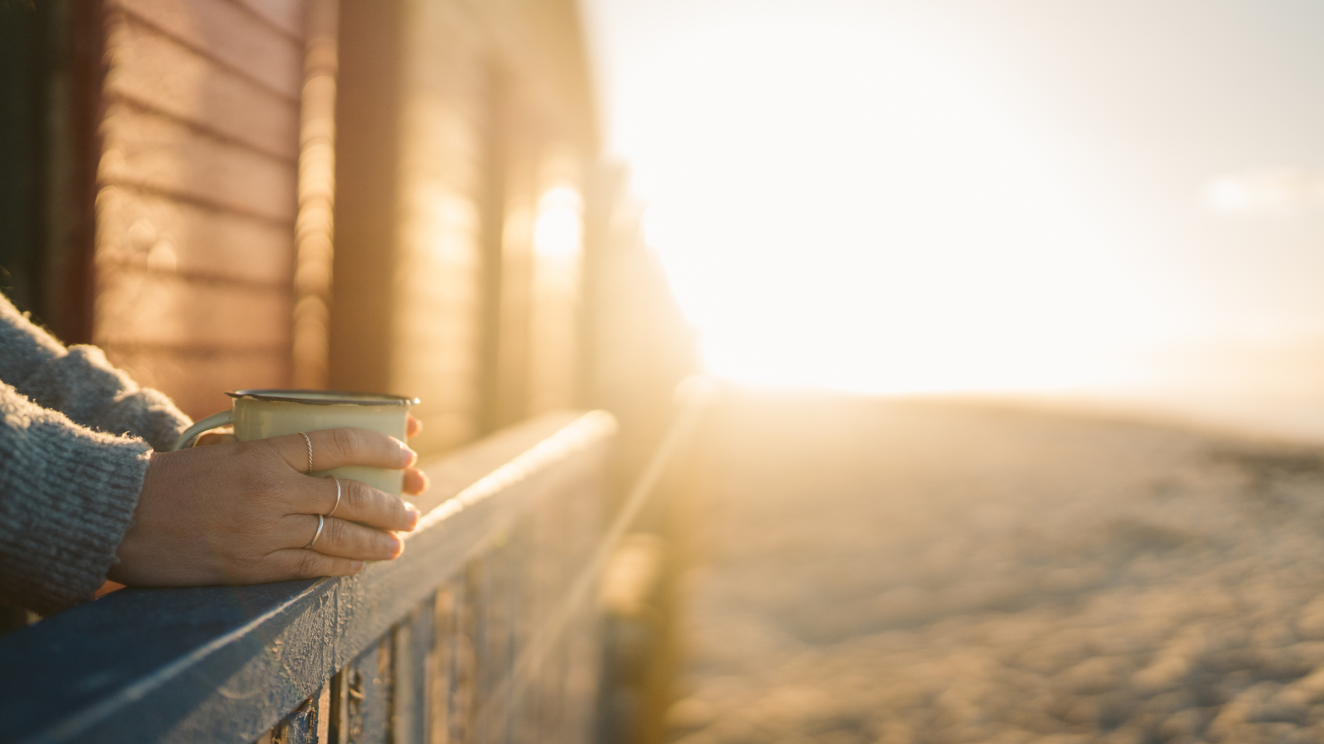 Hands in a knit sweater holding a mug on a wooden balcony overlooking a beach at sunset.