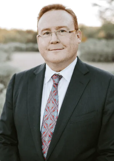 Man in a suit and tie, smiling, outdoors in a soft-focus setting.