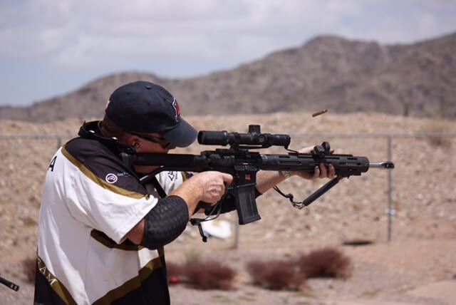 Person shooting a rifle with scope at an outdoor range, mountains in the background, shell casing ejected.