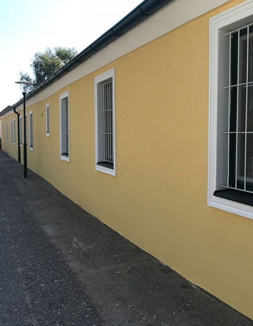 Gelbe Gebäudewand mit weiß umrandeten Fenstern entlang eines schmalen gepflasterten Gehwegs