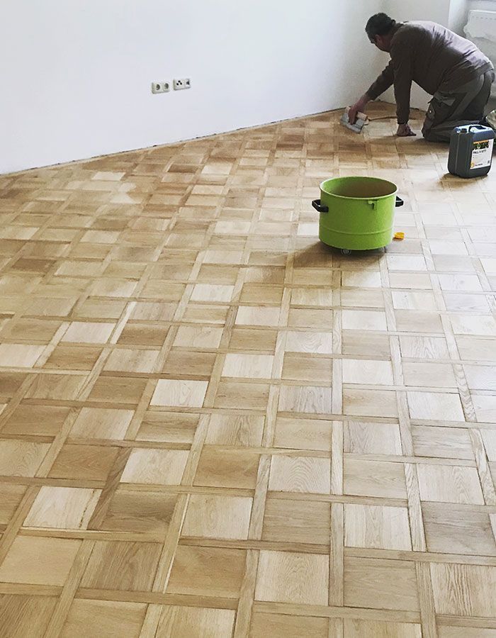 Arbeiter trägt Oberflächenbehandlung auf einen Holzparkettboden auf; im Vordergrund ist ein grüner Eimer zu sehen.