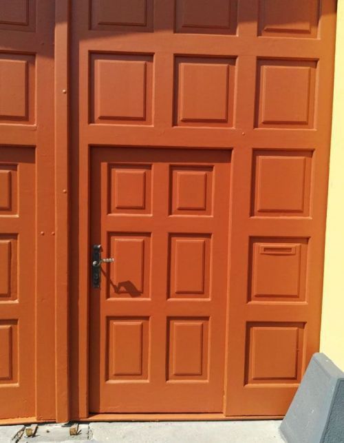 Orangefarbene Doppelflügeltüren mit schwarzem Griff und Schloss vor heller Wand und hellem Boden.