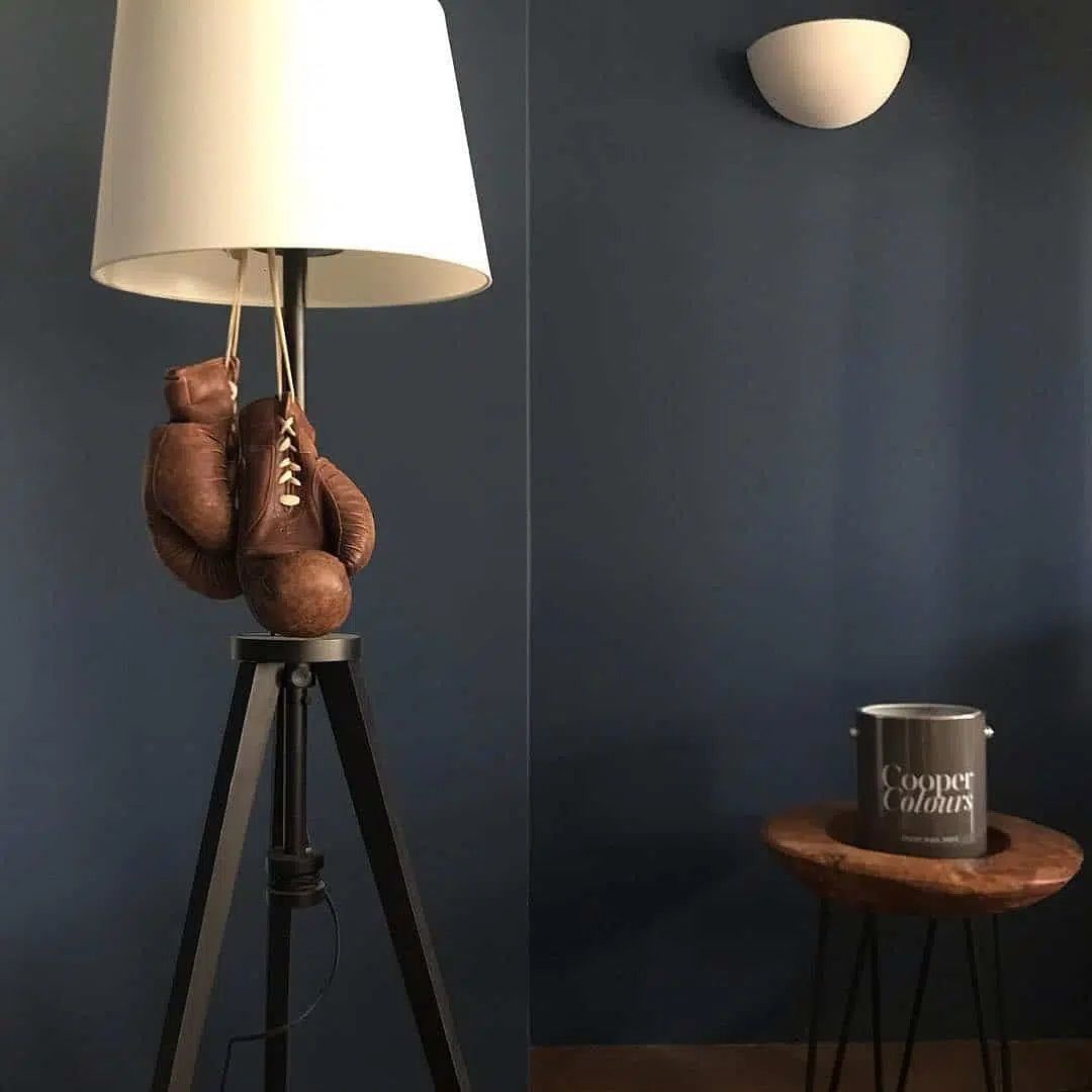Lampe mit weißem Schirm auf einem Stativ neben einem kleinen runden Holztisch vor dunkler Wand