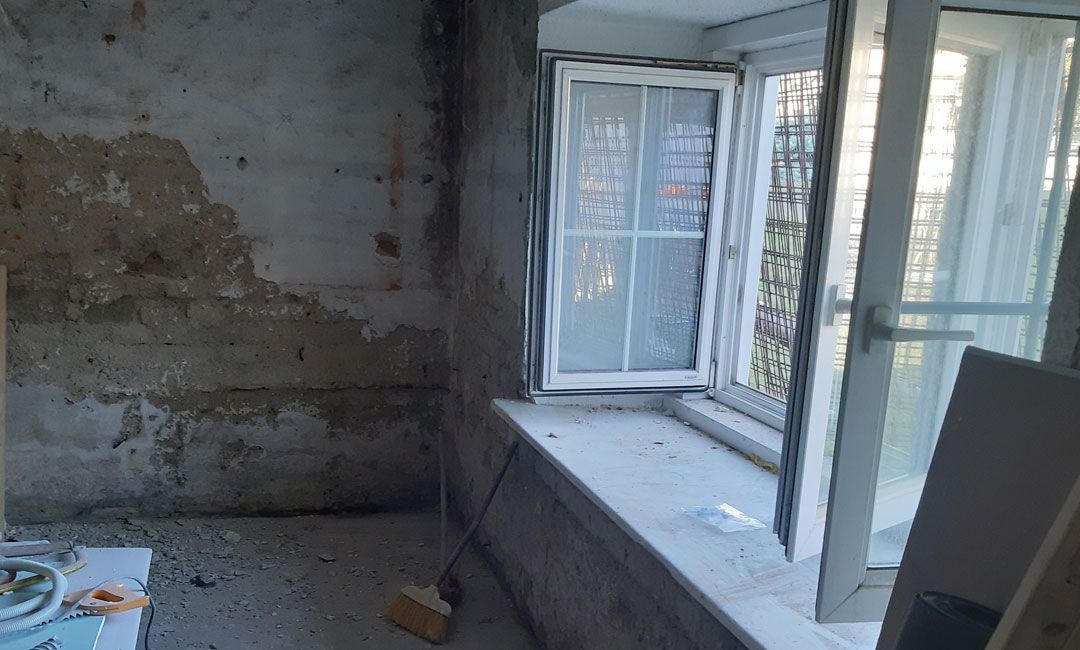 Ein kahler, unfertiger Raum mit Betonwand und einem weiß gerahmten Fenster, das Tageslicht hereinlässt.