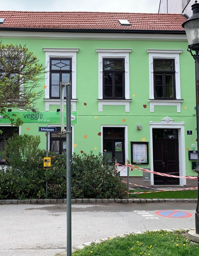 Mintgrünes Gebäude mit weißen Zierelementen, rotem Dach und Schaufenstern hinter einer Straßenlaterne und Sträuchern.