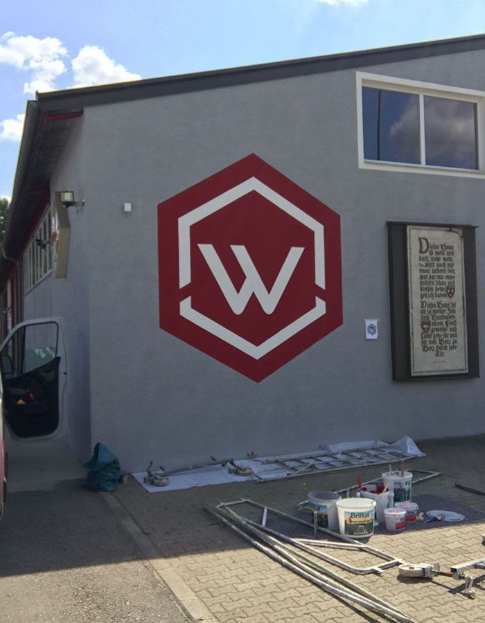 Graue Gebäudewand mit einem roten Sechseck-Logo und einem weißen „W“, Werkzeuge und Materialien liegen auf dem Boden.