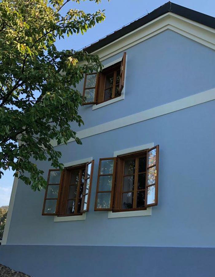 Blaue Hausfassade mit braunen Fensterläden und weißen Zierelementen, von unten neben einem belaubten Baum gesehen.