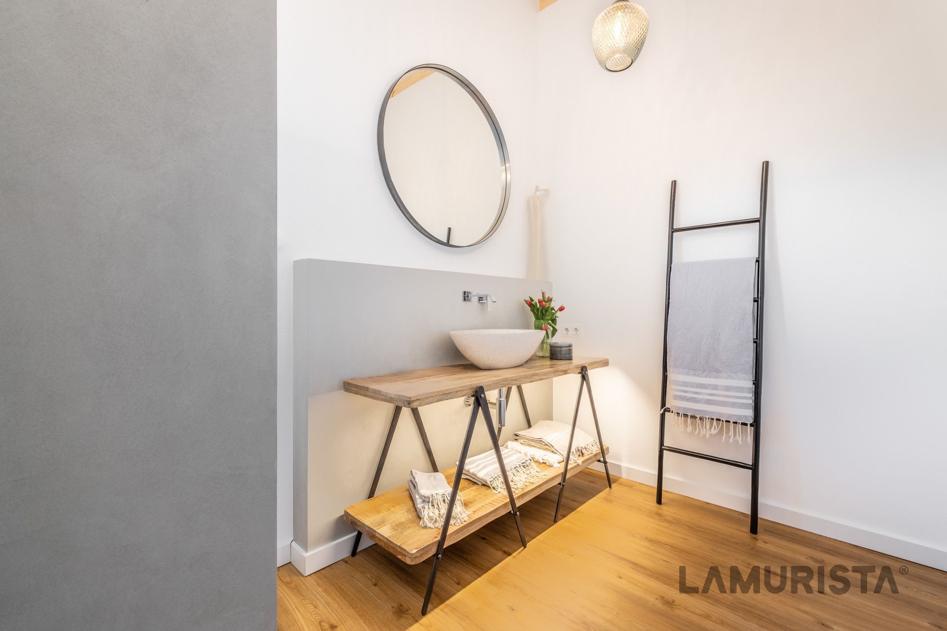 Minimalistische Badezimmerecke mit rundem Spiegel, Holzkonsole, Handtuchhalter in Leiterform und warmem Licht über Fischgrätparkett.