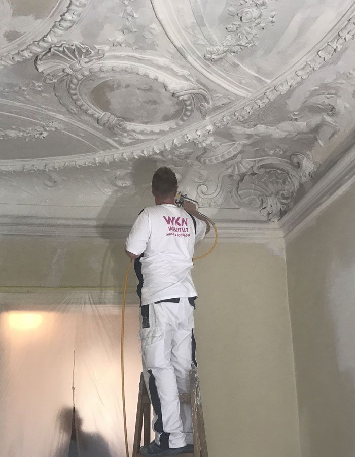 Ein Arbeiter streicht auf einer Leiter die verzierte weiße Decke in einem Raum mit beigen Wänden.