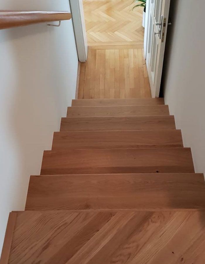 Eine Holztreppe führt hinab in einen Flur mit hellen Wänden und einer offenen Tür am unteren Ende.