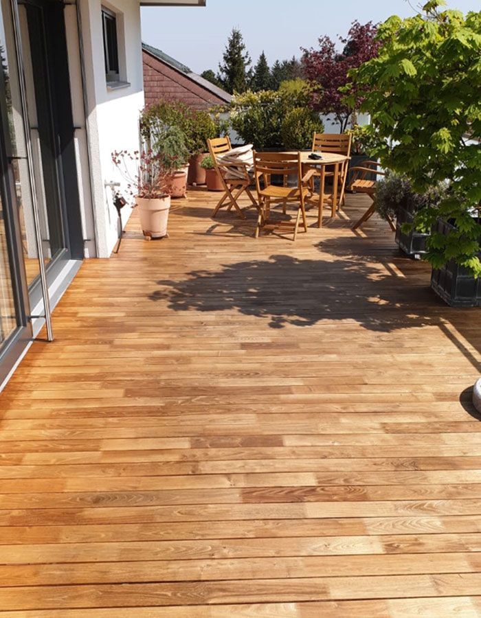 Holzterrasse auf dem Dach mit Gartentisch, Stühlen, Topfpflanzen und Blick auf Bäume und Nachbarhäuser.
