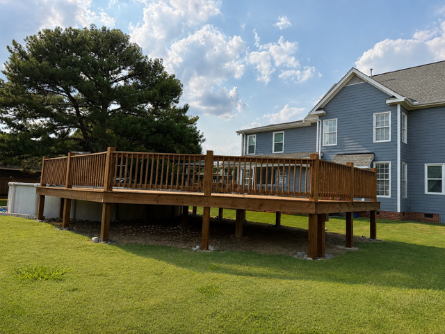 Wooden deck with railing beside a blue house on a grassy yard, under a partly cloudy sky