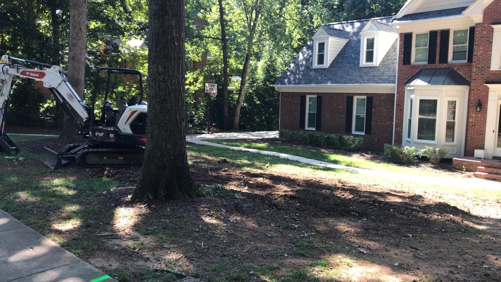 Suburban house with a large tree in the foreground and an excavator parked in the driveway.