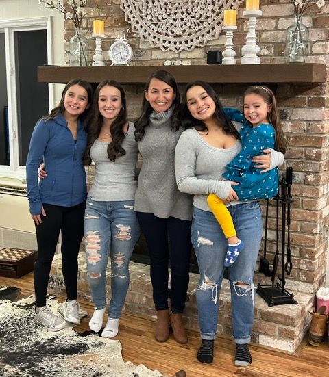 Five women posing indoors in front of a brick fireplace, smiling and standing close together.