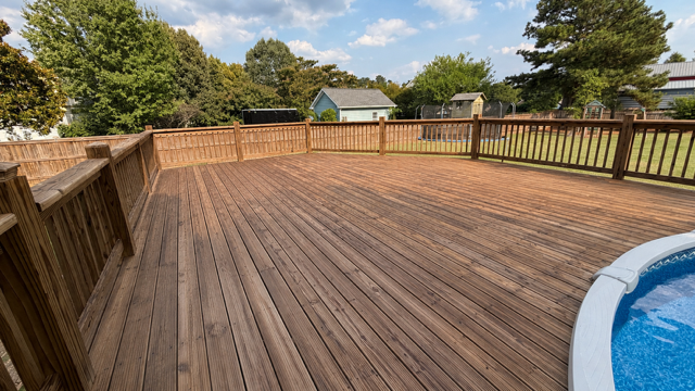Spacious wooden deck with railing, trees beyond, and a blue pool at the edge