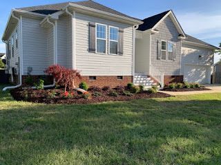 Suburban single-story beige house with a front lawn, white shutters, and landscaped shrubs