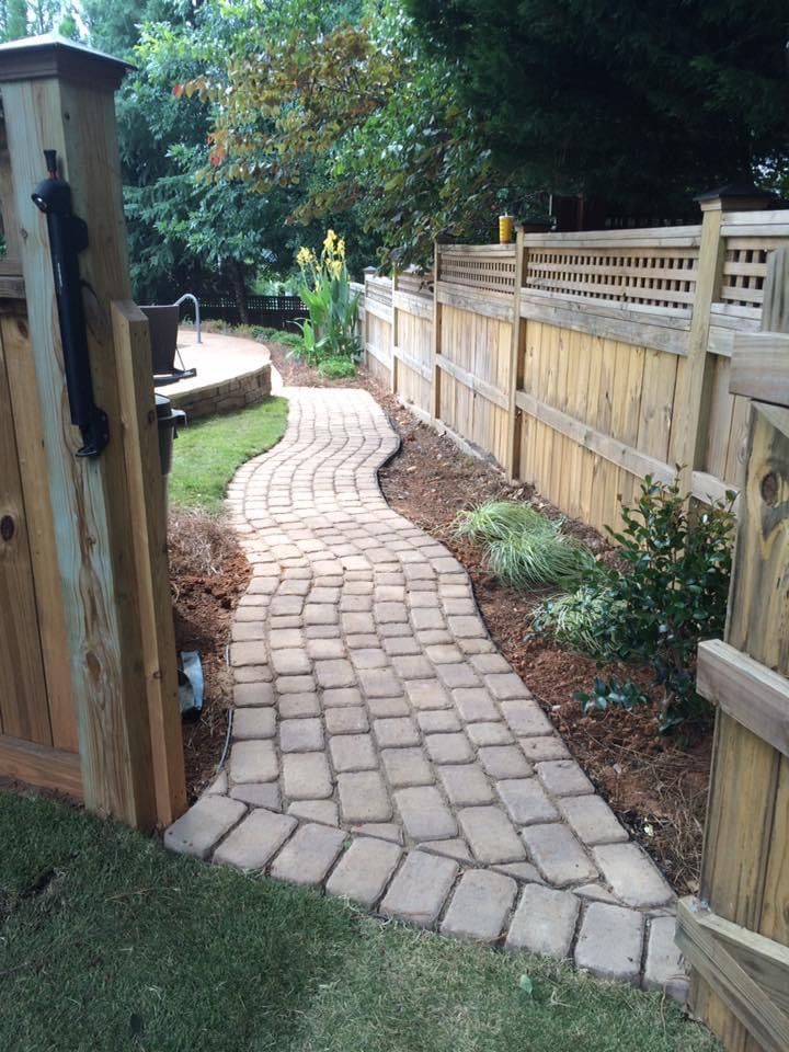 Curved brick walkway through a landscaped yard with a wooden fence and gate opening to a garden.